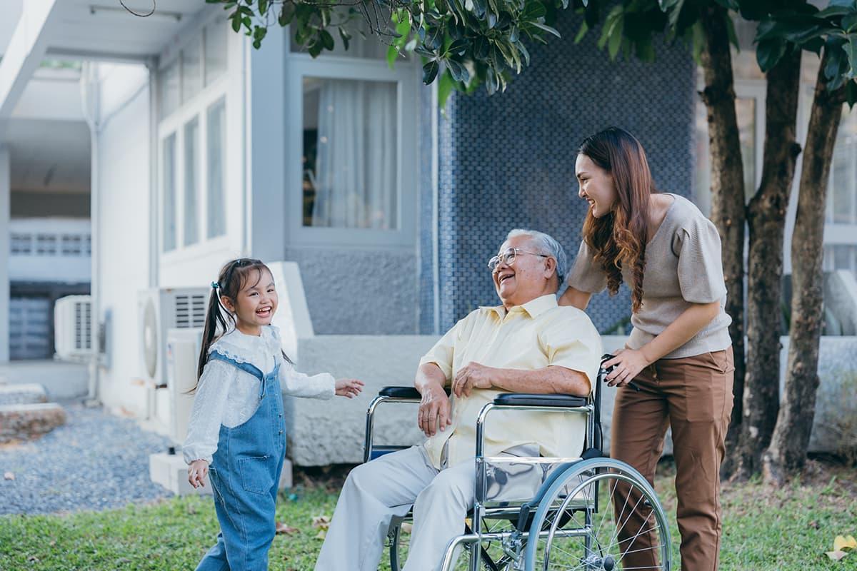 一名社工与一位坐轮椅的老人和他的孙女在树下的房子外面一起笑.